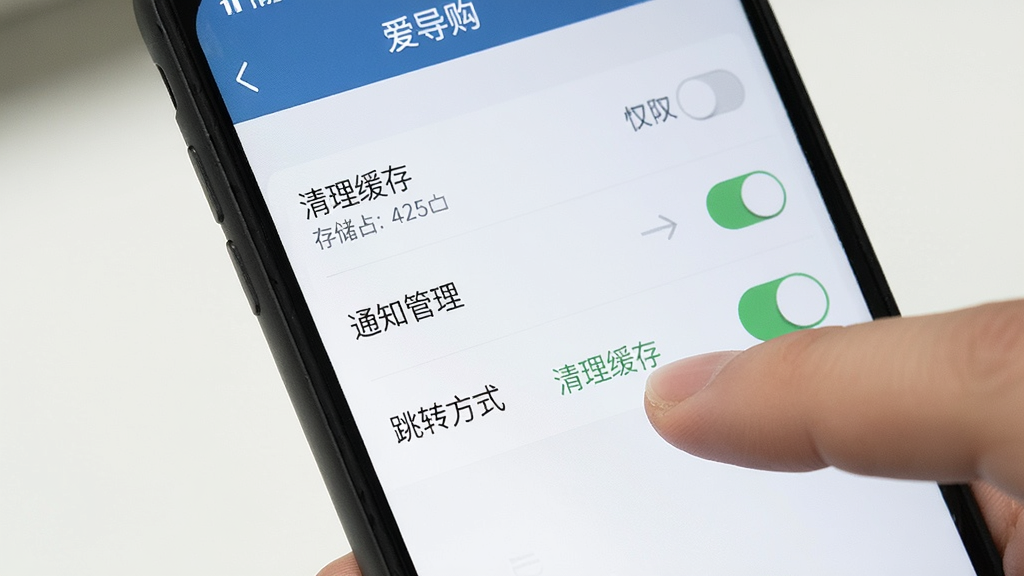 手机屏幕显示“爱导购”设置页面,包含“清理缓存”“通知管理”“跳转方式”等选项,旁边有一只手正点击“清理缓存”按钮;布局为特写构图,风格为简洁工具类界面,细节包括存储占用数字和权限开关,画质清晰,画面干净,无多余元素