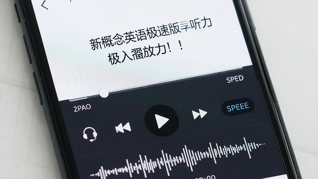 一张新概念英语极速版听力播放界面的特写图,主体为手机屏幕上的音频播放器,布局居中显示课文标题与进度条,排版包含播放控制按钮与倍速选项,风格简约现代,细节有耳机图标与音频波形,画质清晰,画面干净