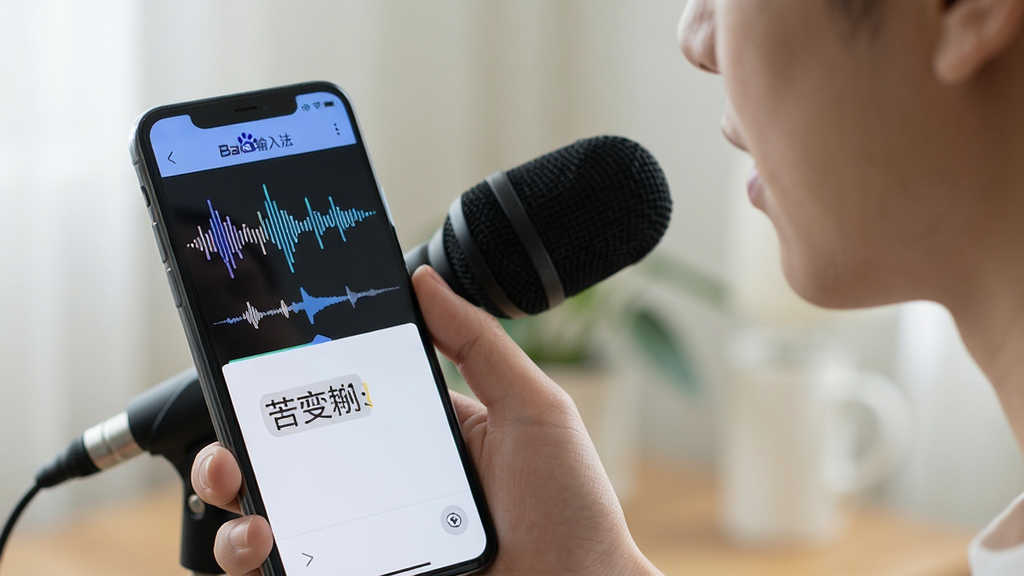 人物手持手机对着麦克风说话,手机屏幕显示“百度输入法”语音输入波形与转写文字,背景虚化,布局简洁,排版突出人机交互,风格自然,细节呈现声波动画与文字同步,画质干净,无多余元素。