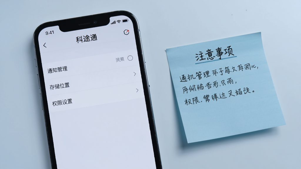 一部手机显示科途通的设置页面,页面中有“通知管理”、“存储位置”、“权限设置”等选项,背景为浅灰色,手机旁边有一张写着“注意事项”的便签纸,整体色调偏冷,清晰干净