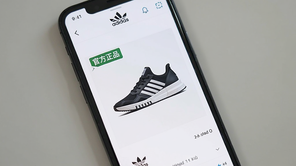 画面主体为一部手机,手机屏幕显示阿迪达斯App的商品详情页,页面中有“adidas”品牌标识和一双运动鞋的展示图,旁边有“官方正品”字样标签,布局简洁,风格清新,画质干净,无多余元素。