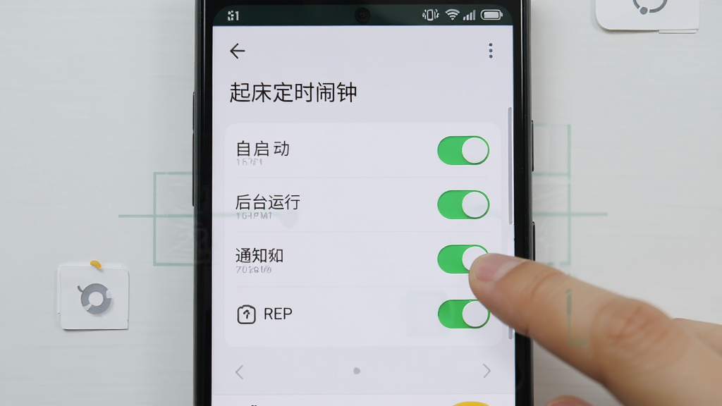 一部安卓手机屏幕显示“起床定时闹钟”的权限管理页面,页面上有自启动、后台运行、通知等多个权限开关,旁边有手指正在点击开关,背景是简洁的浅灰色桌面,风格为操作指引图,细节包括开关状态高亮、文字清晰、图标简约,画质高清,干净,无多余元素